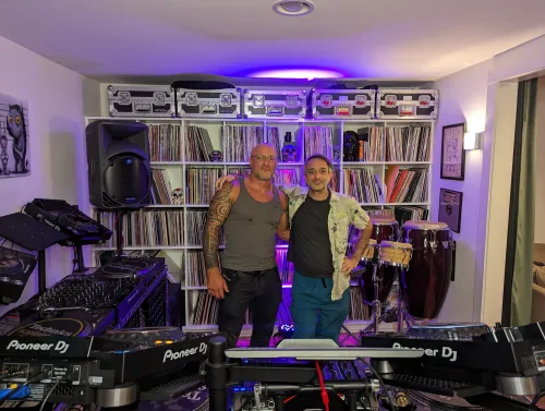 Phil Anthony and Pezzner posing behind Pioneer DJ equipment in a home studio with extensive vinyl record collection and purple ambient lighting