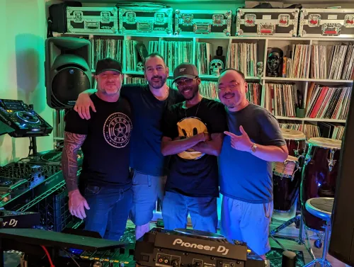 Phil Anthony, Paul Velocity, Tony H and sangdo posing together in a home studio with Pioneer DJ equipment, vinyl record collection, and green ambient lighting