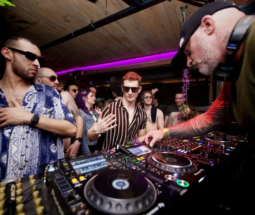 DJ performing on a beach at sunset with a laptop and controller setup under a white tent