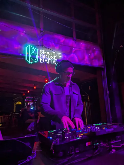 DJ mixing beneath the Seattle House Mafia neon sign under purple stage lighting