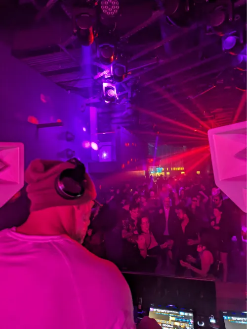 View from behind the DJ booth across a packed dance floor bathed in red and magenta light